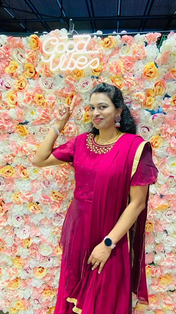 SaiSudha is wearing a pink dress and standing in front of a wall covered in rose blossoms, pointing her finger towards a neon text saying 