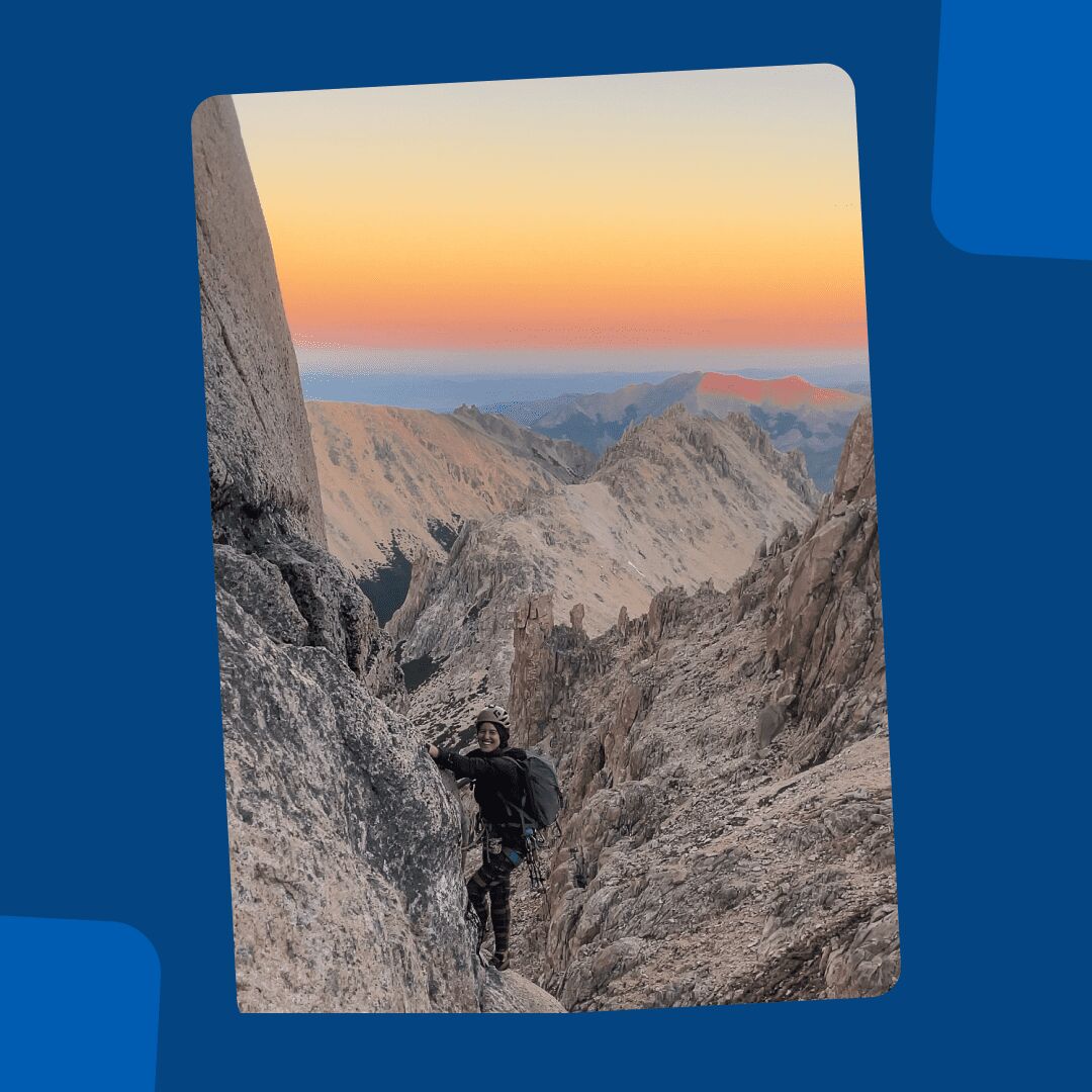 Delfina climbing a cliff, a mountain range in the background and the sky is colored yellow and red from sunrise.