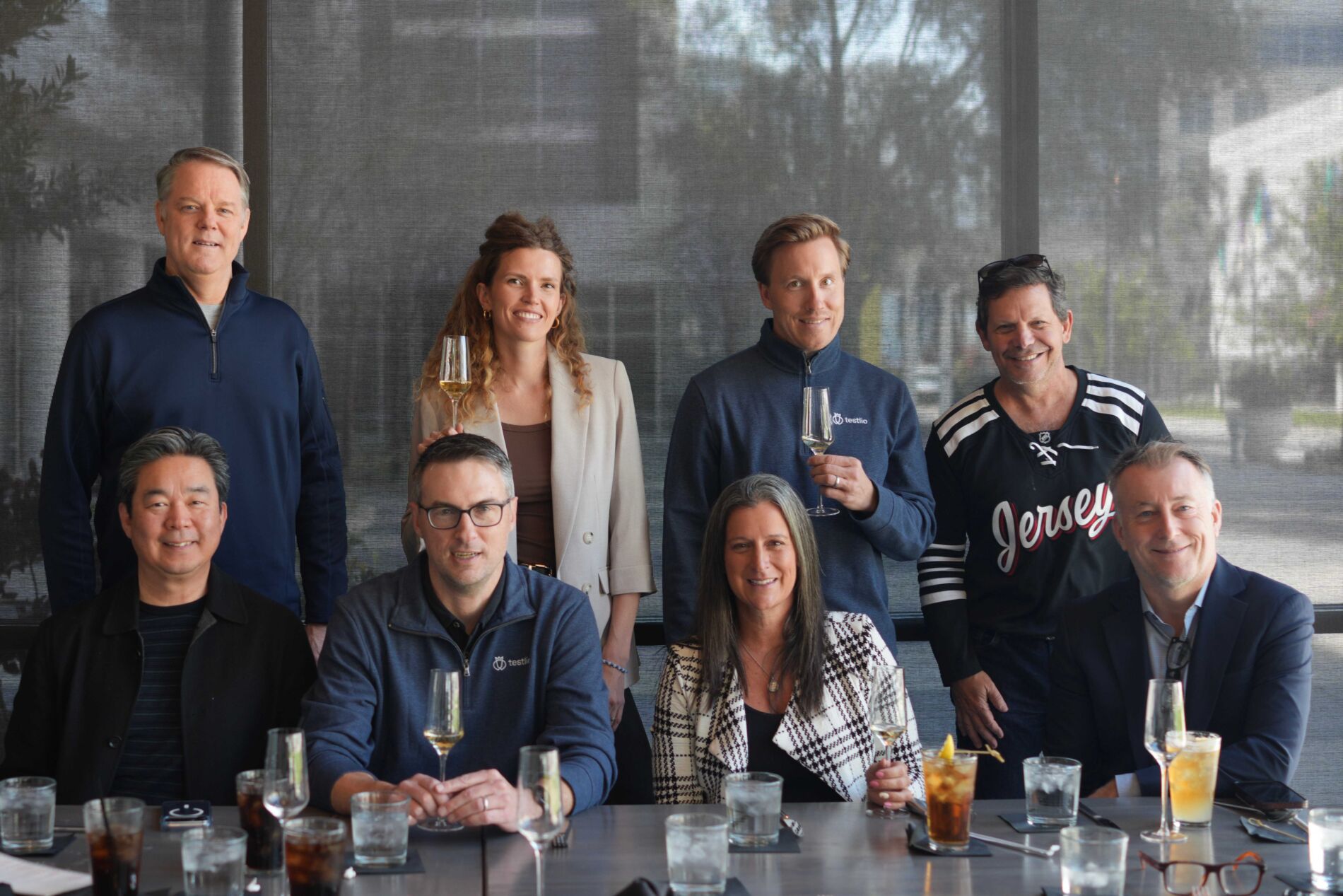 Group photo of Testlio’s board members and executive leadership team seated and standing around a restaurant table, some holding wine glasses. The group is arranged in two rows, with four people seated in front and four standing behind. From left to right, bottom row: Insik Rhee (Board Director), Mike Stearns (VP of Marketing), Summer Weisberg (CEO), and Dean Hickman-Smith (CRO). From left to right, top row: Brian Benson (CFO), Kristel Kruustük (Founder and Board Director), Marko Kruustük (Co-founder and Board Director), and Dave Keil (Board Director). Not pictured: Carmen Scarpa (Board Director) and Anthony Lee (Board Observer).