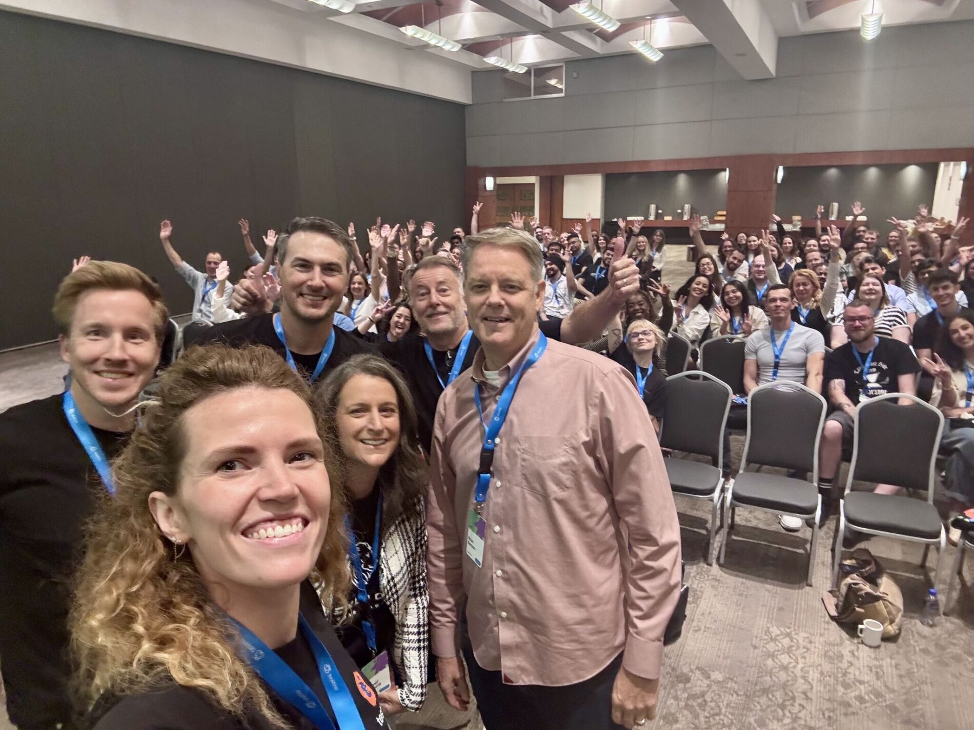 A large group of people from Testlio pose together in a conference room during LionFest. The photo is taken as a cheerful selfie from the front row, showing smiling team members and many people in the background waving and raising their hands.